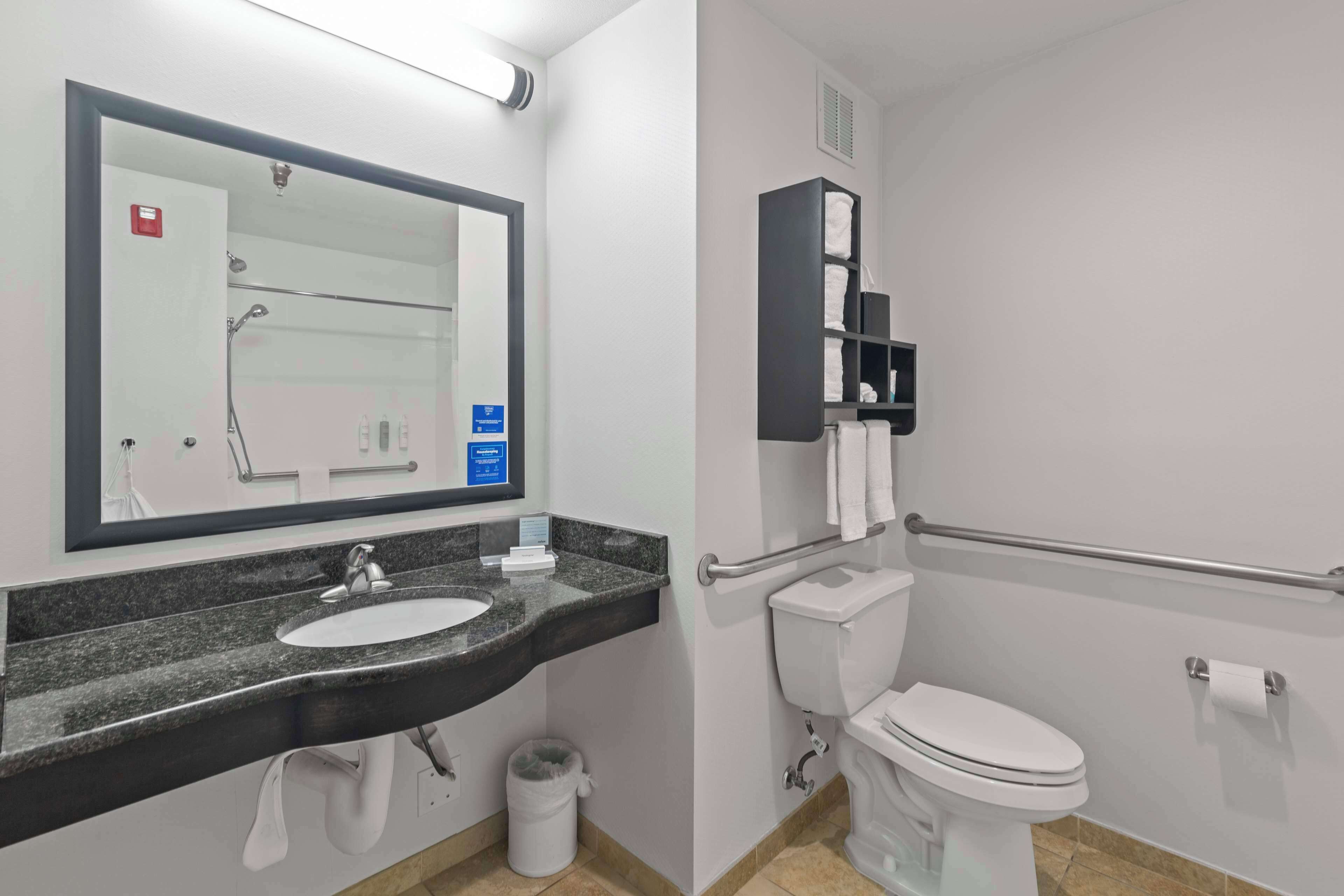 Un baño moderno con lavamanos de granito, un espejo, inodoro y barandilla para accesibilidad.