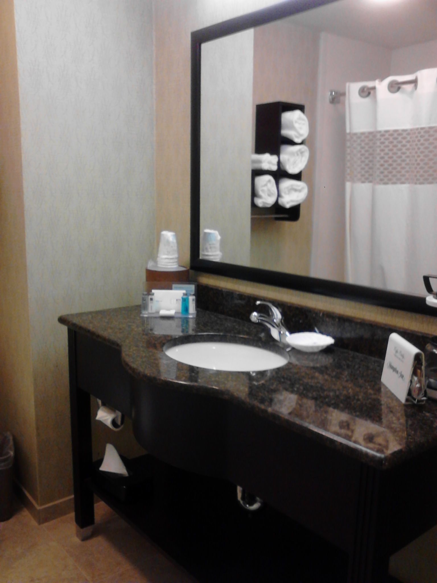 A hotel bathroom with a granite countertop sink, a mirror, and a towel rack with towels.