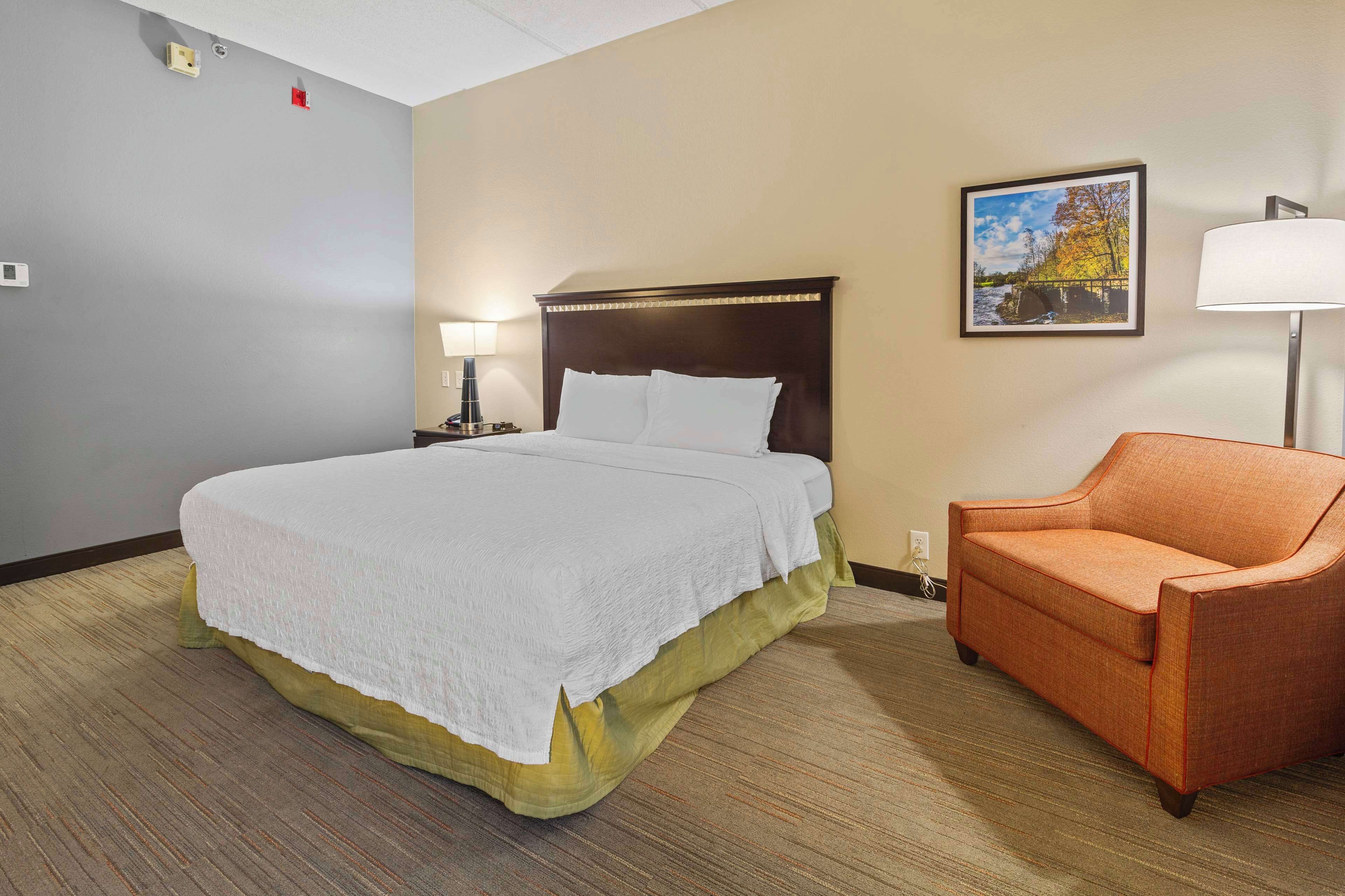 A hotel room with a large bed, a nightstand with a lamp, a chair, and a painting on the wall.