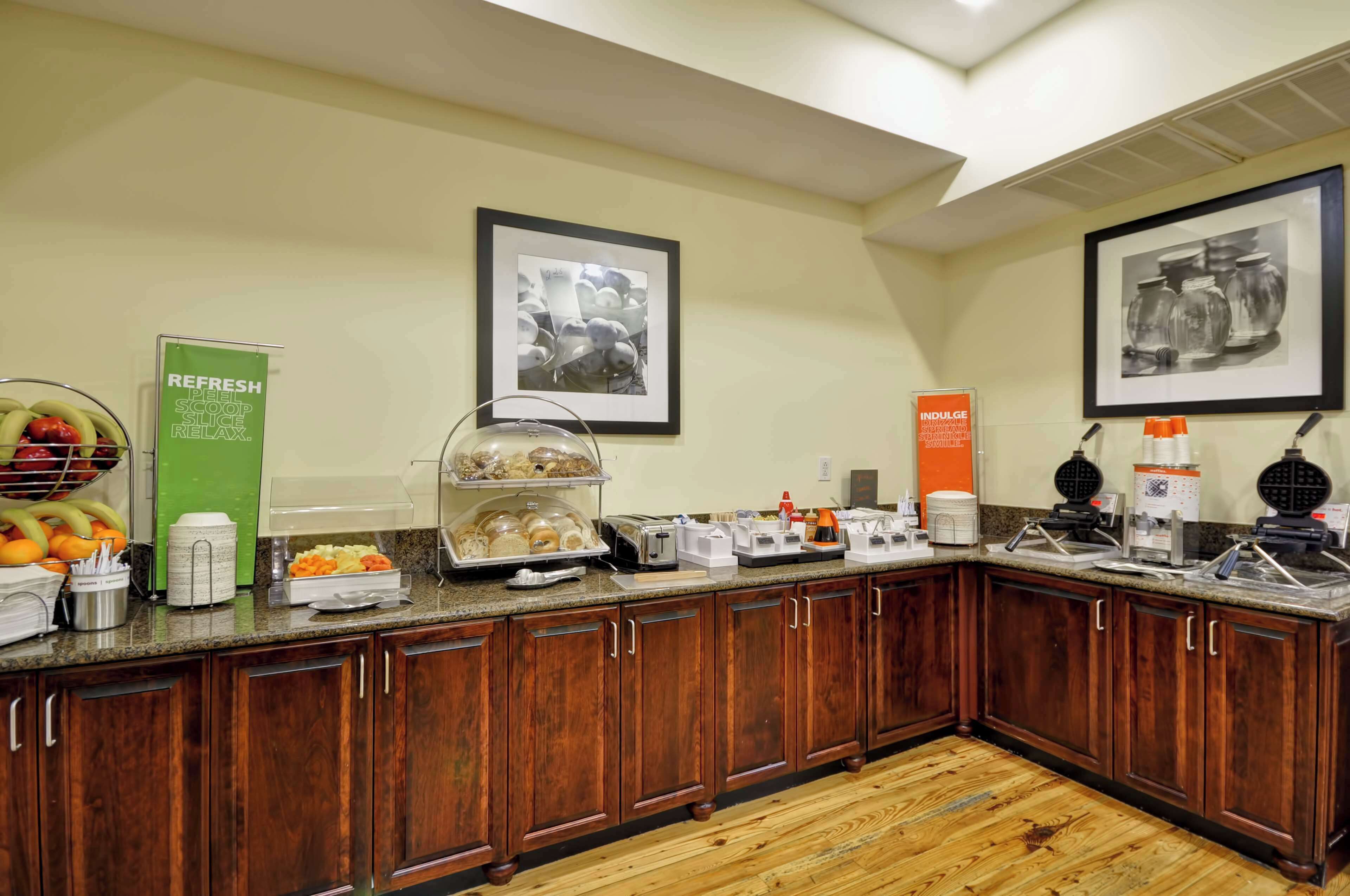 A hotel kitchen with wooden cabinets, a fruit bowl, a coffee station, and framed pictures on the wall.