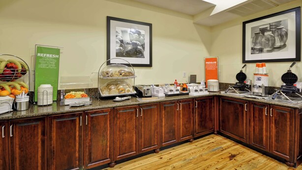 A hotel kitchen with wooden cabinets, a fruit bowl, a coffee station, and framed pictures on the wall. - Hampton Inn & Suites Charleston/West Ashley (Charleston)