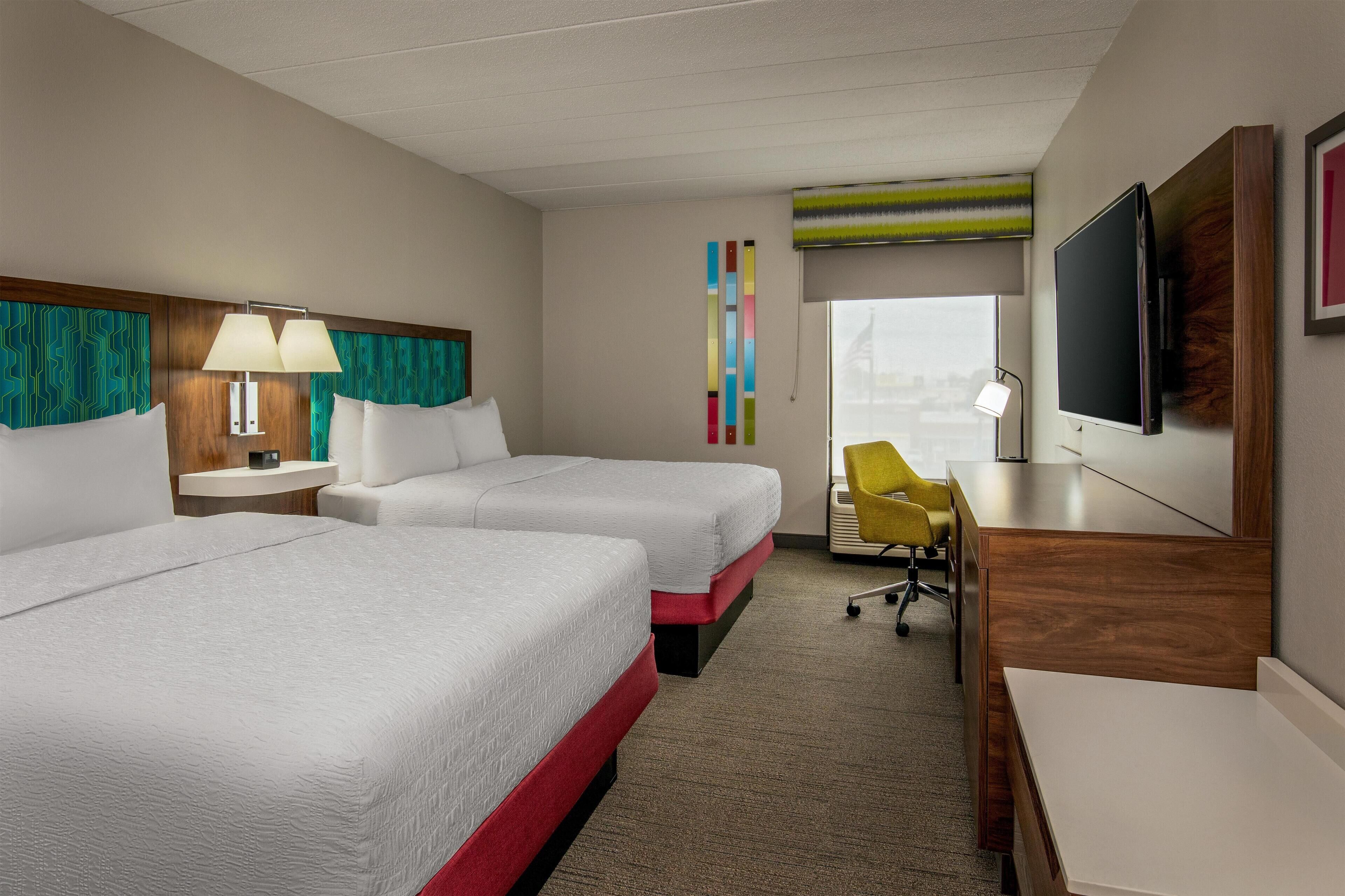 A hotel room with two beds, a desk, a chair, a TV, and a window.