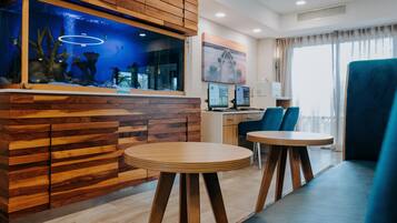 A modern hotel lobby with a wooden reception desk, a large aquarium, two round tables, and a seating area with blue chairs.