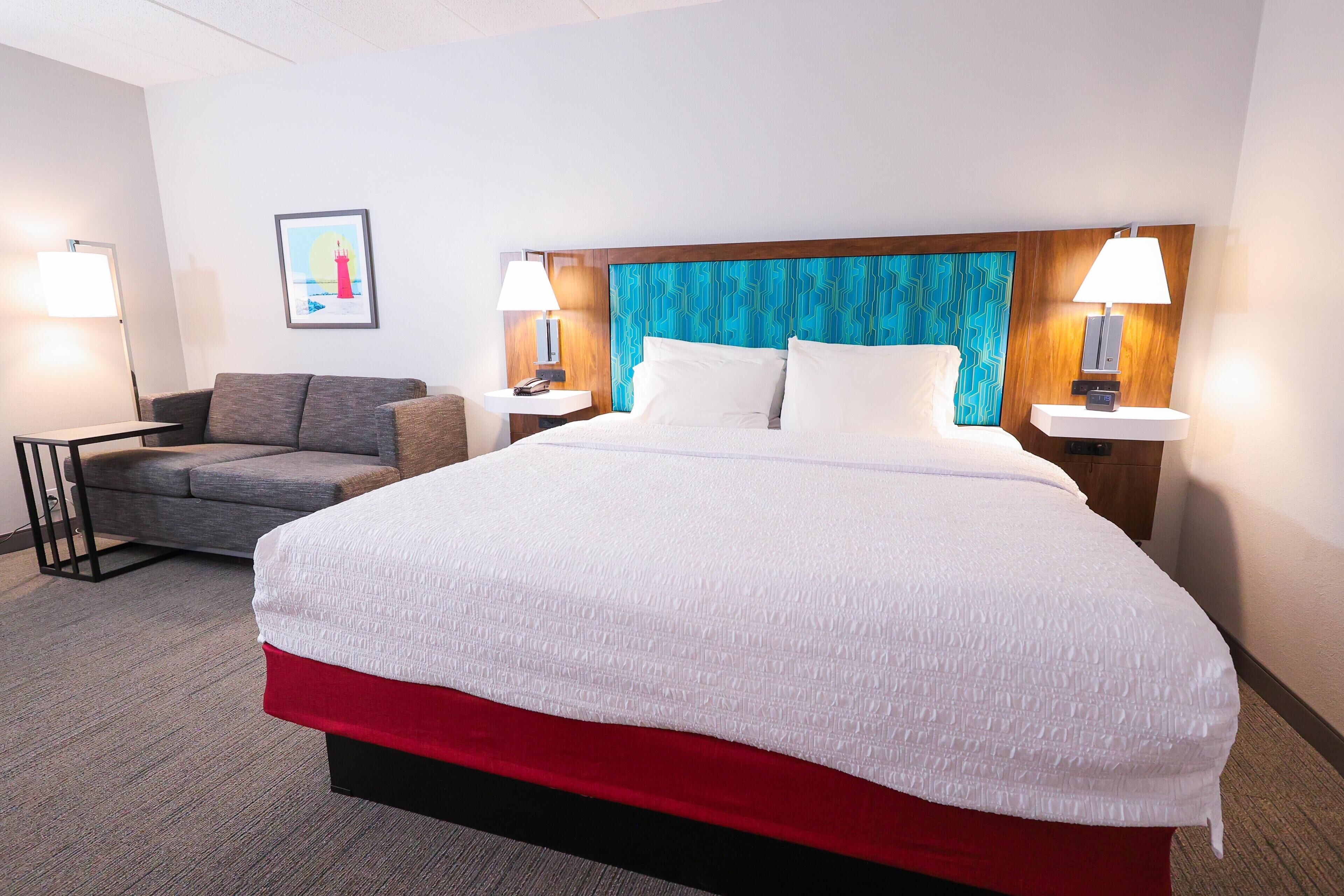 A hotel room with a bed, a sofa, two bedside lamps, a framed picture on the wall, and a carpeted floor.