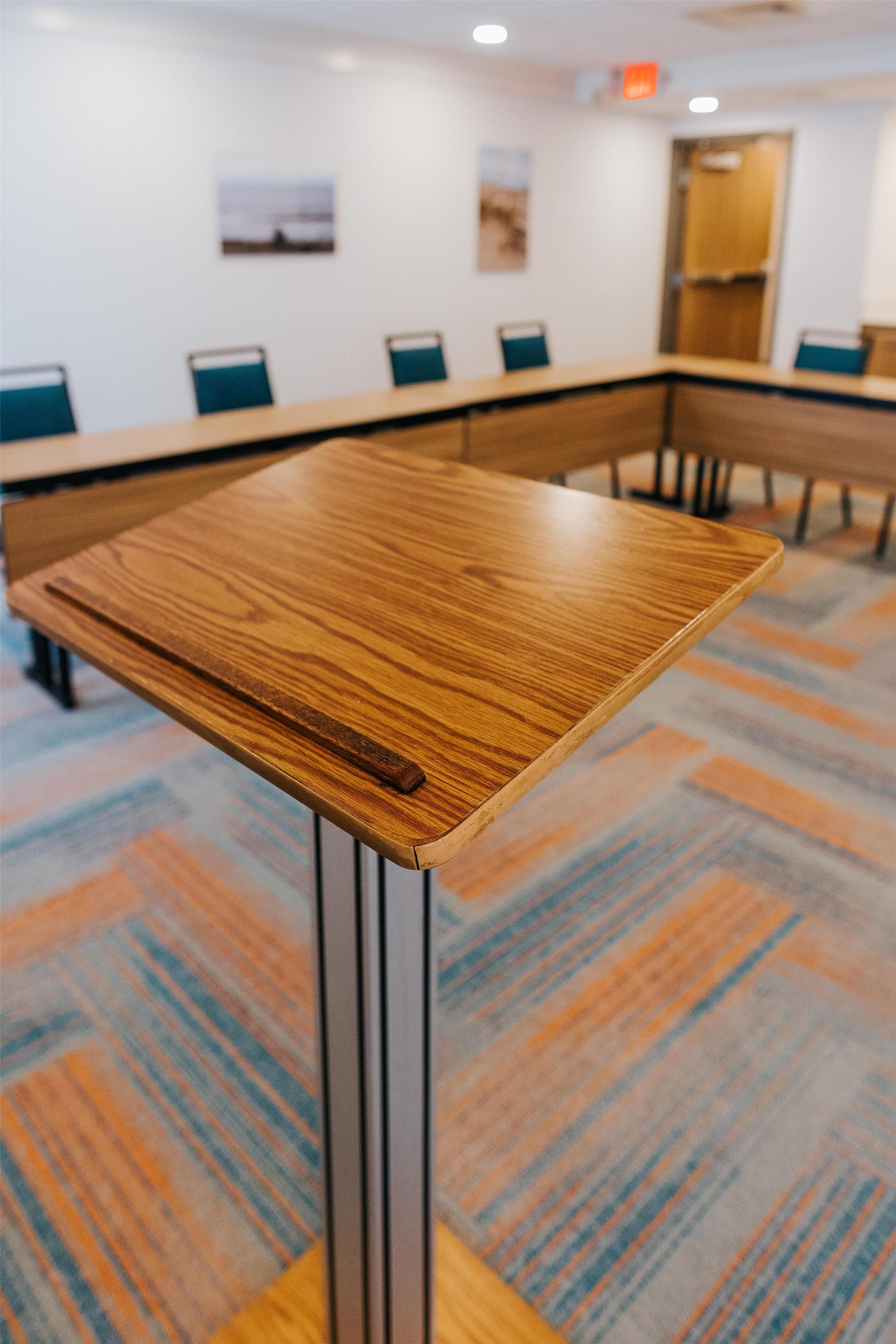 A conference table with a wooden top and metal legs, surrounded by chairs with blue cushions, in a room with patterned carpet and framed pictures on the wall.
