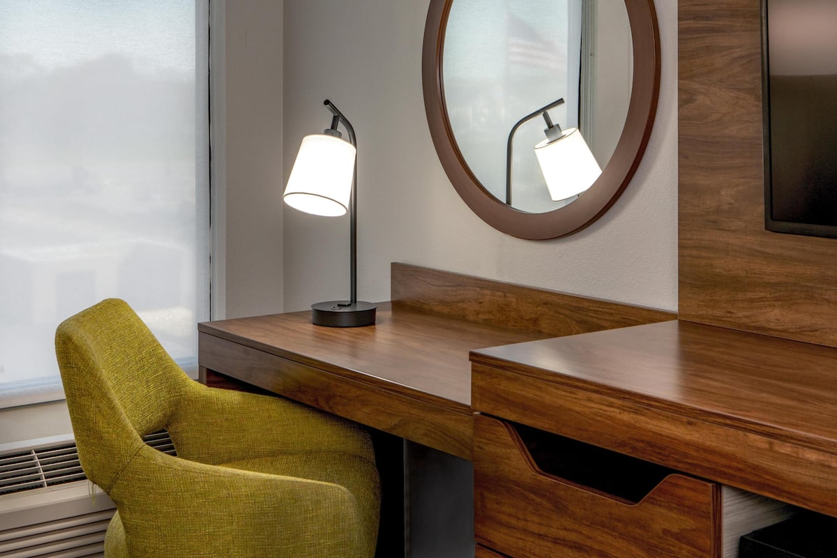 A wooden desk with a lamp, a mirror, and a green chair in a hotel room.