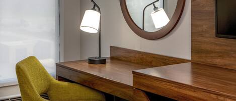 A wooden desk with a lamp, a mirror, and a green chair in a hotel room.