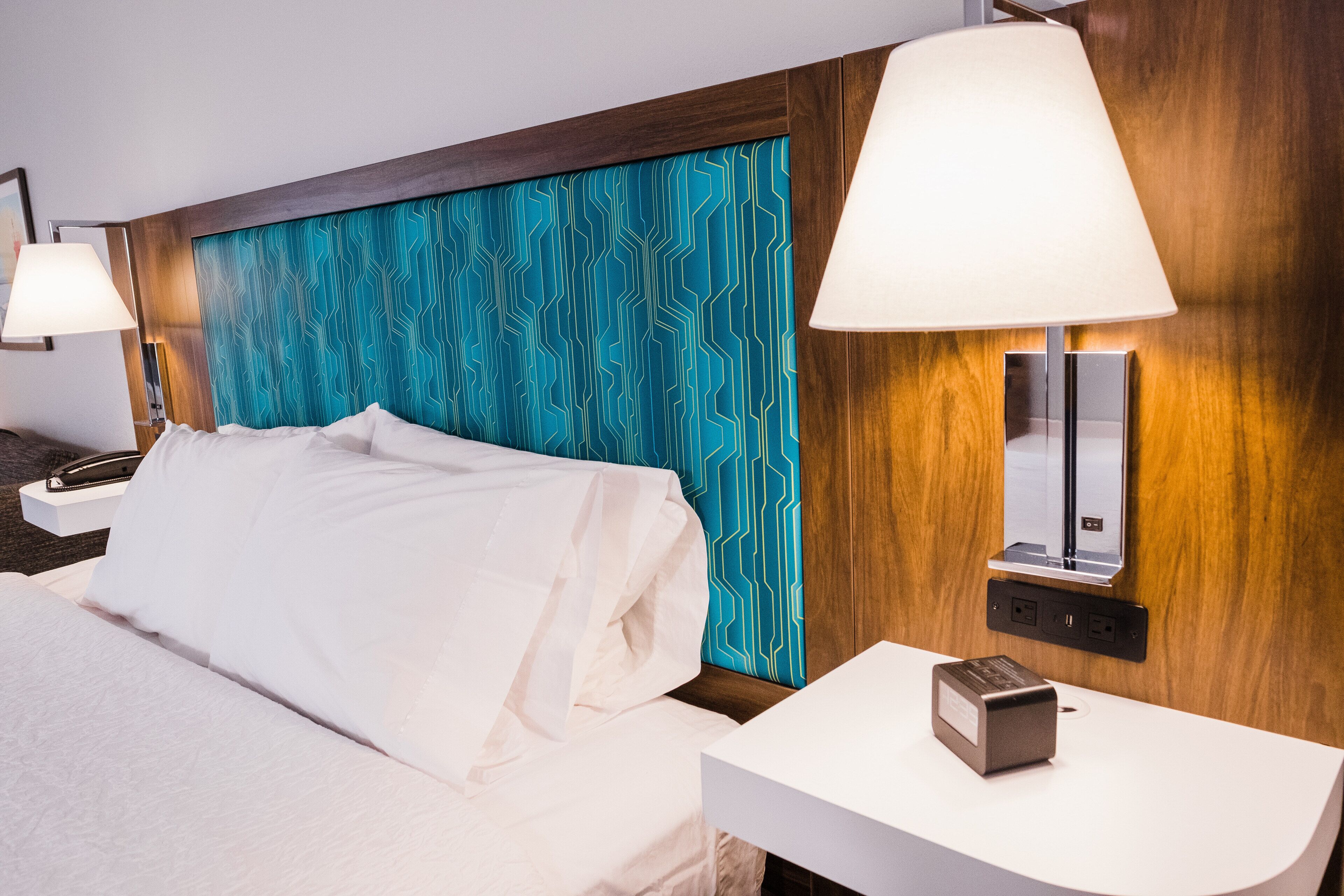 Habitación de hotel con cama, mesita de noche, lámpara y estación de carga en la pared.