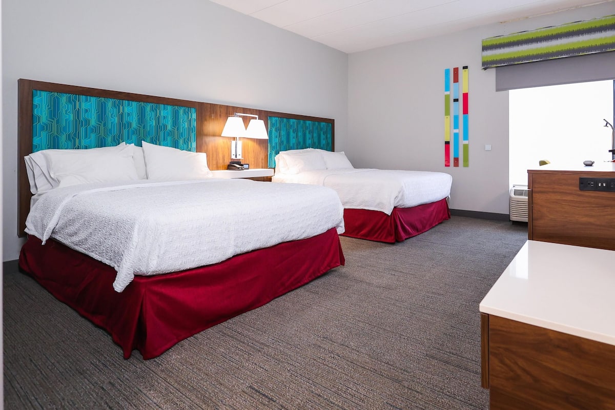 A hotel room with two beds, a desk, a lamp, and a colorful wall decoration.