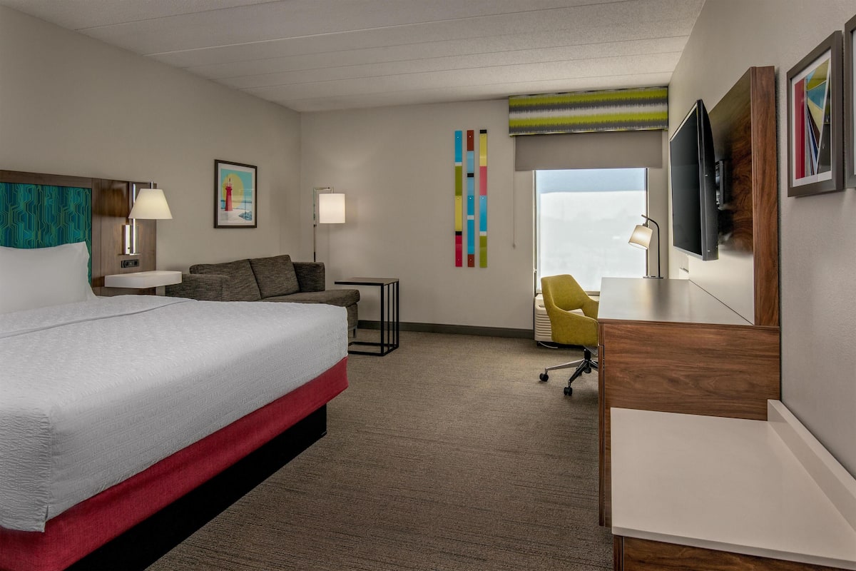 A hotel room with a bed, a desk, a chair, a TV, and a window.