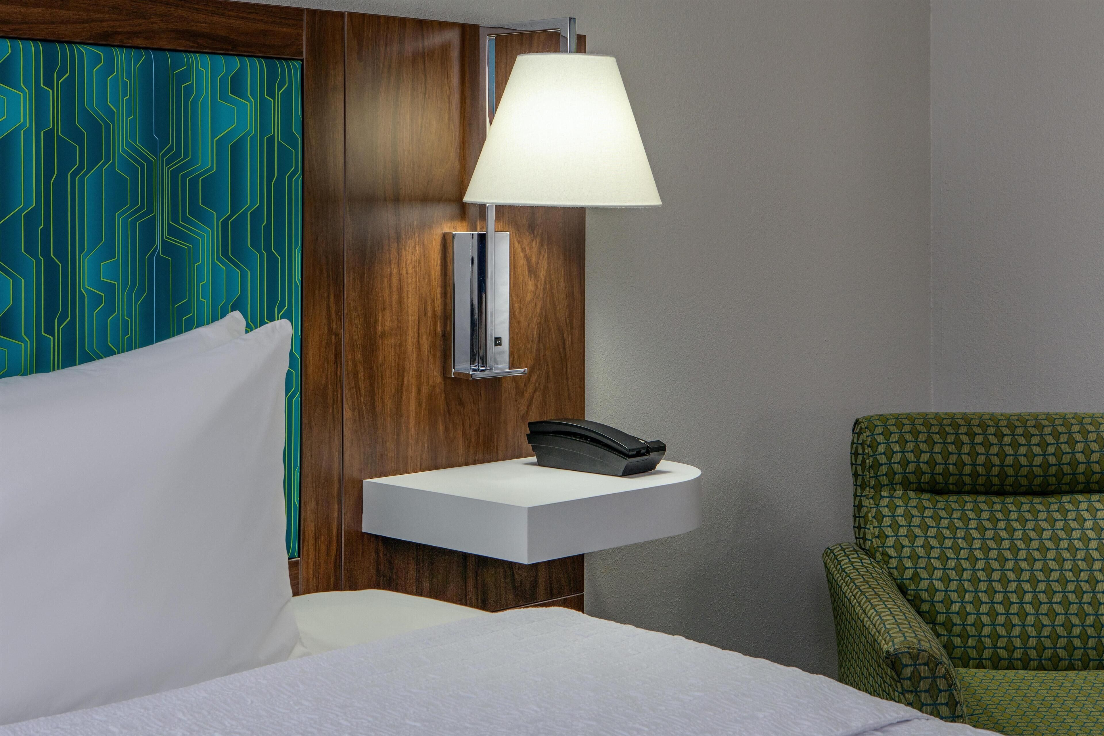 Een hotelkamer met een bed, een nachtkastje met een lamp, een telefoon en een groene fauteuil met een patroon.