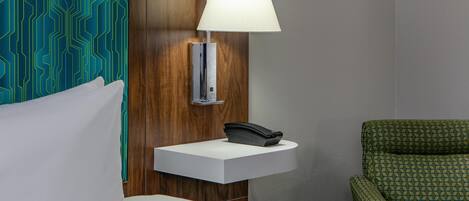 Een hotelkamer met een bed, een nachtkastje met een lamp, een telefoon en een groene fauteuil met een patroon.