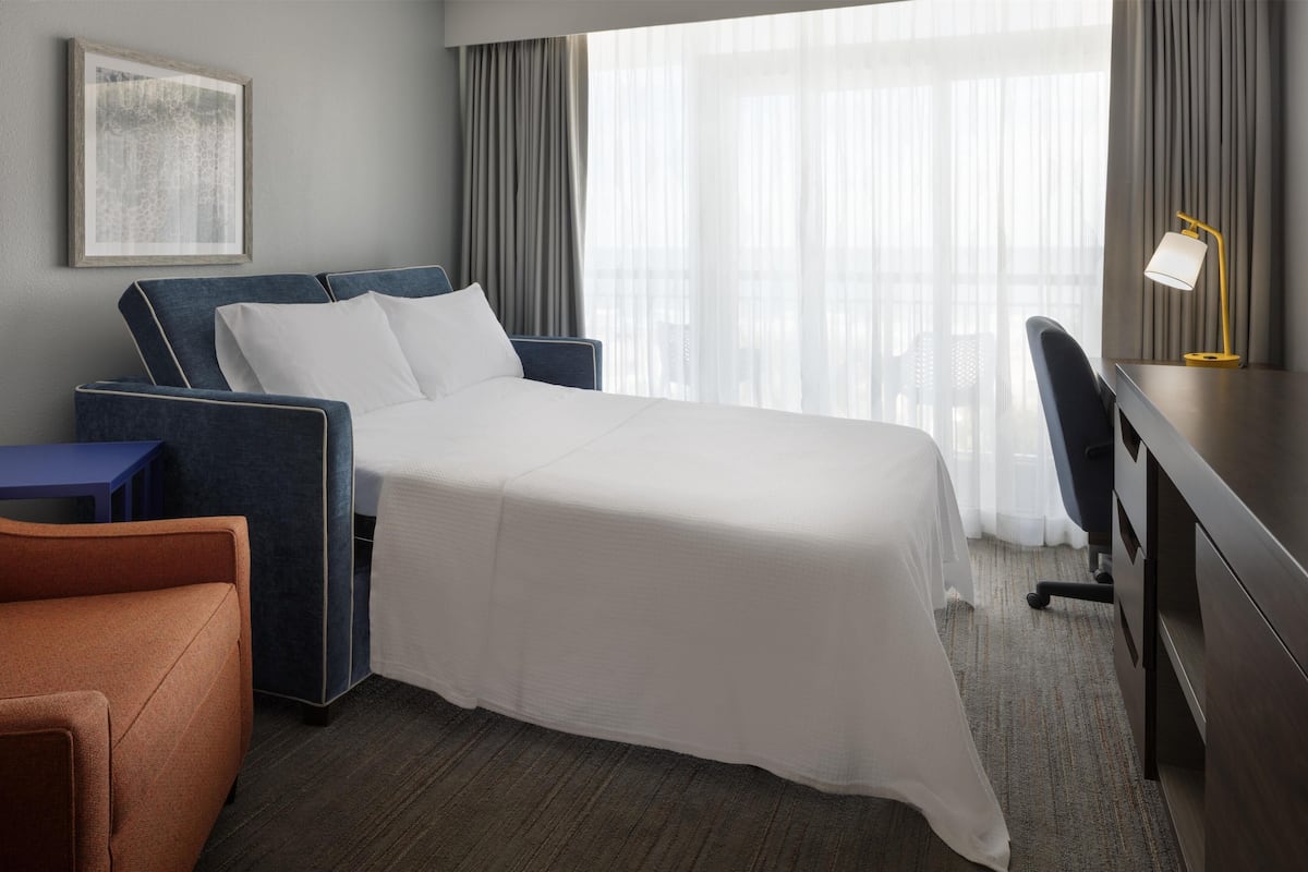 A hotel room with a bed, a desk, a chair, and a large window with curtains.