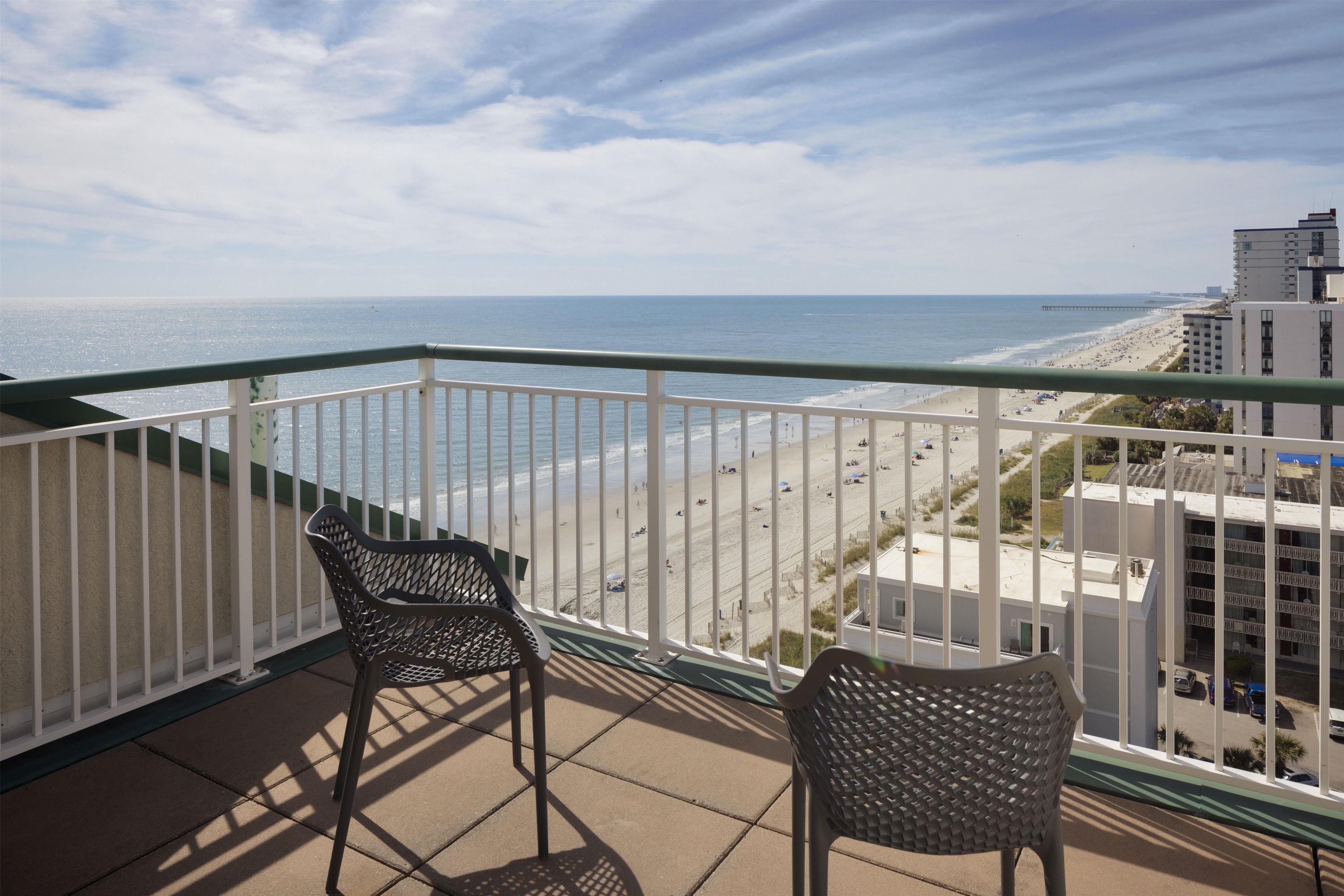 A balcony with two chairs overlooking a beach with a pier in the distance.