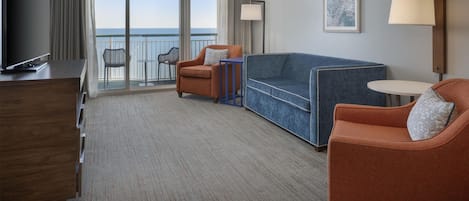 A hotel room with a television, a sofa, two armchairs, a small table, a lamp, and a view of the ocean through large windows.