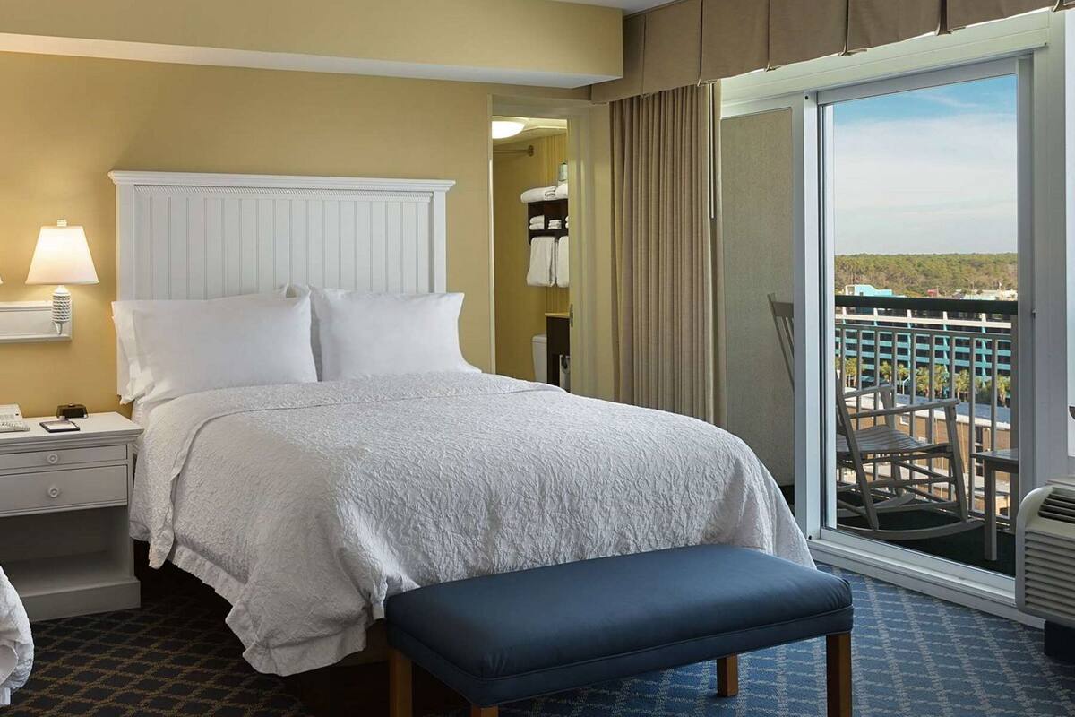 A hotel room with a bed, a bench, a nightstand, a lamp, a window with a view, and a door.