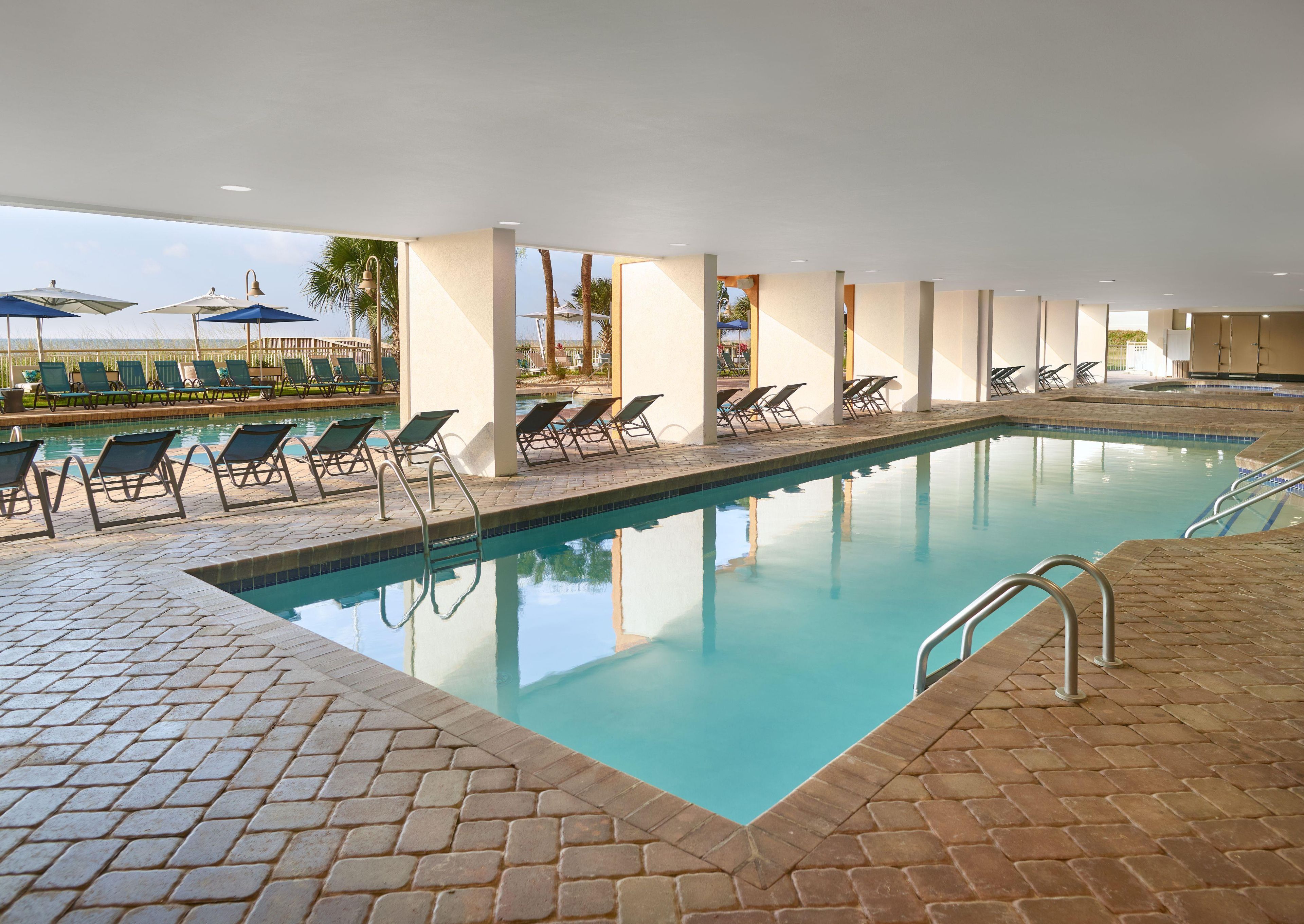 An indoor swimming pool with lounge chairs and umbrellas, surrounded by a tiled deck.