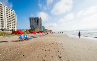 Una playa con filas de sillas de playa azules y rojas, un puesto de socorrista y un alto edificio de hotel al fondo.