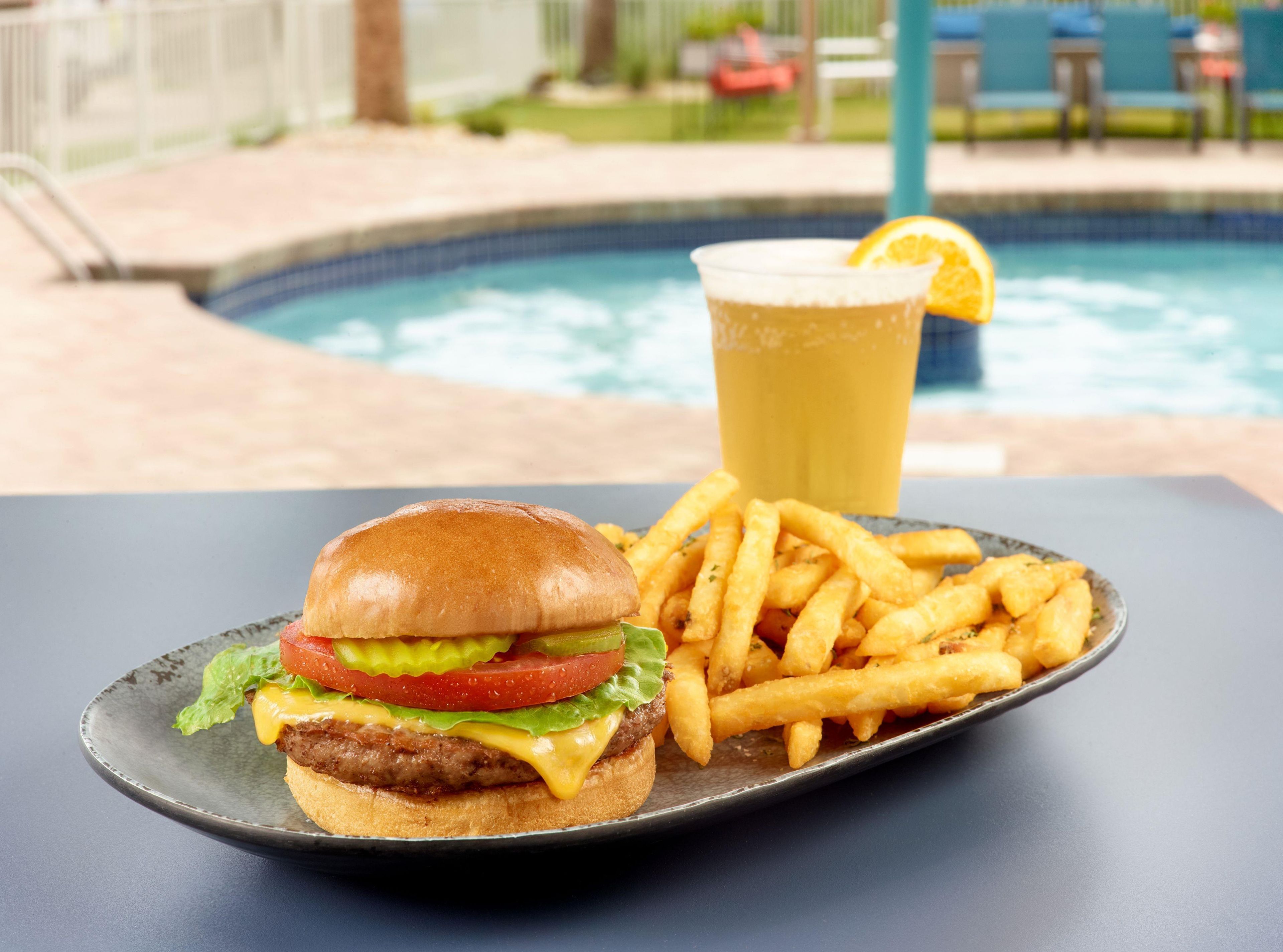 Un repas composé d’un hamburger, de frites et d’un verre de bière, servi sur une table au bord d’une piscine.