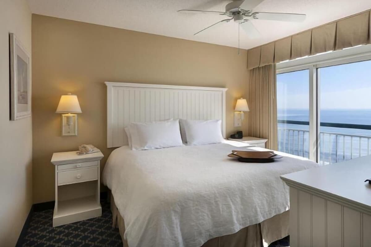 A hotel room with a bed, bedside lamps, a nightstand, a desk with a chair, a ceiling fan, and a view of the ocean.