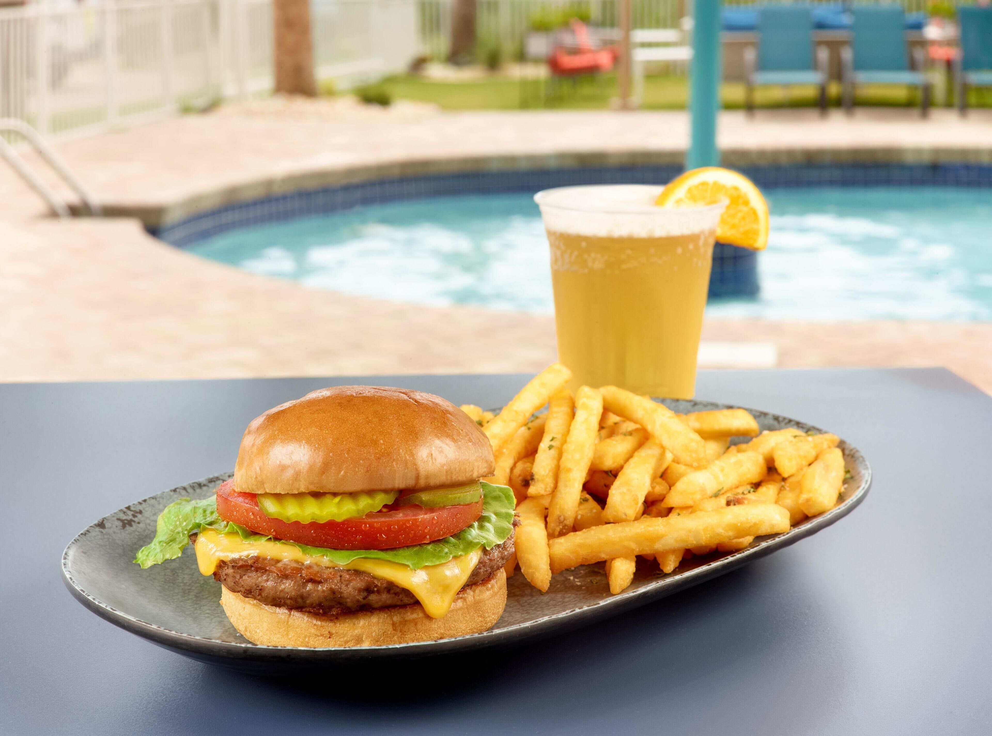 Un repas composé d’un hamburger, de frites et d’un verre de bière, servi sur une table au bord d’une piscine.