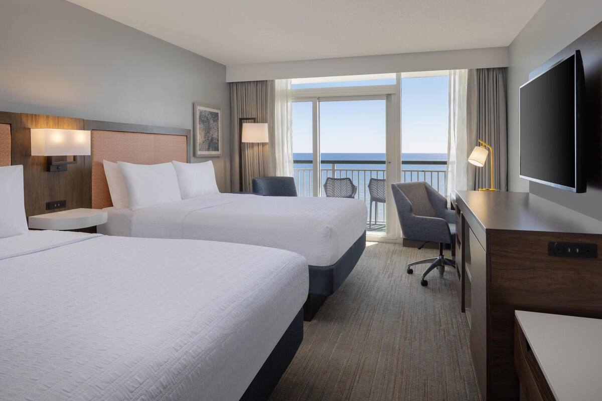 A hotel room with two beds, a desk, a chair, a TV, and a balcony with a view of the sea.