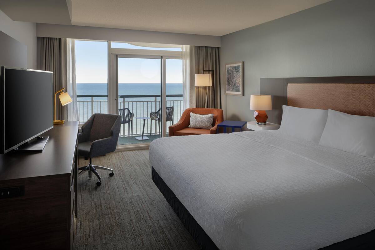 A hotel room with a large bed, a desk with a TV, a chair, a small table with a lamp, and a balcony with a view of the sea.