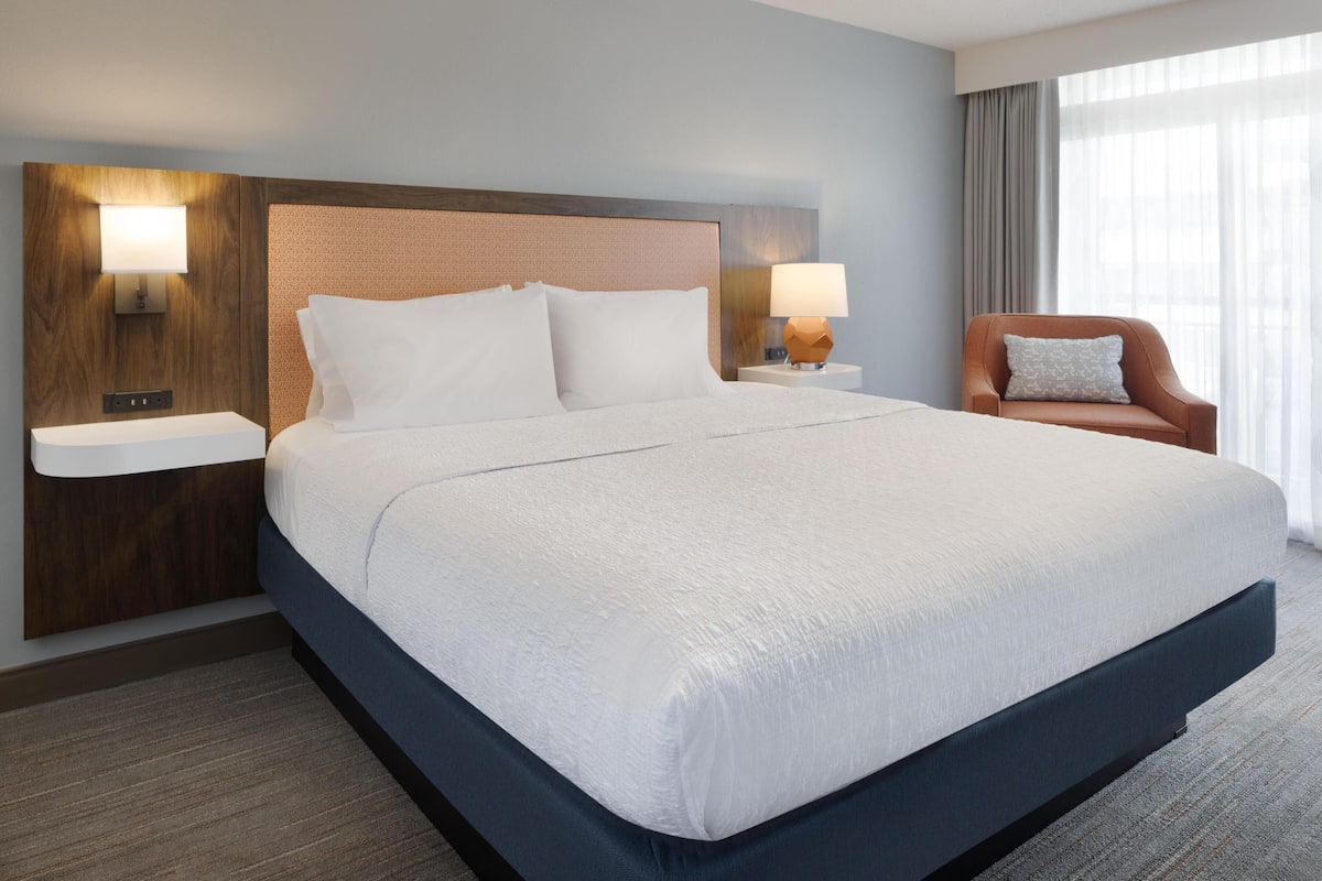 A hotel room with a large bed, two bedside tables, a nightstand, a chair, and a window with curtains.