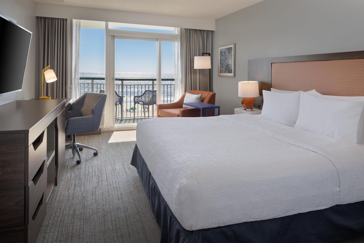 A hotel room with a large bed, a desk, a chair, a small table, and a balcony with a view of the sea.