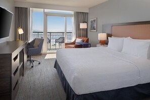 A hotel room with a large bed, a desk, a chair, a small table, and a balcony with a view of the sea.