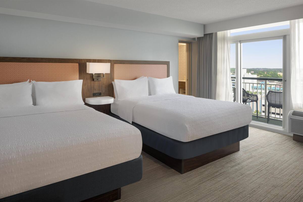 A hotel room with two beds, a nightstand, a lamp, a window with a view, and a balcony with chairs.