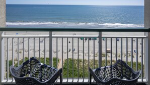 A balcony with two wicker chairs, overlooking a beach with people and a clear sky.
