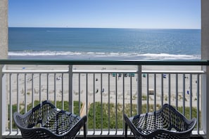 A balcony with two wicker chairs, overlooking a beach with people and a clear sky.