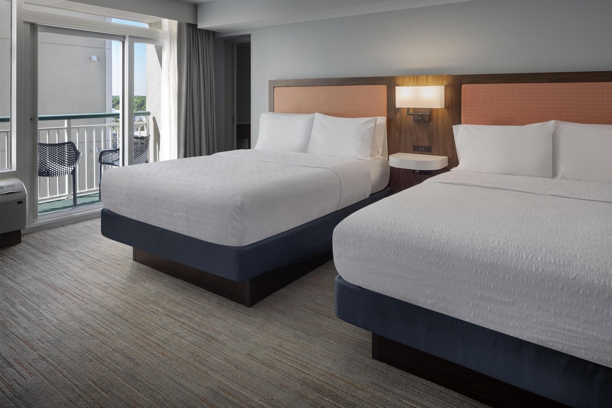 A hotel room with two beds, a balcony with chairs, and a nightstand with a lamp.