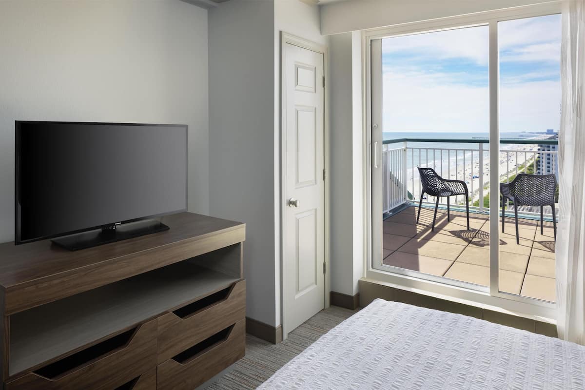 A hotel room with a TV on a wooden stand, a bed, a sliding glass door to a balcony with chairs, and a view of the beach.