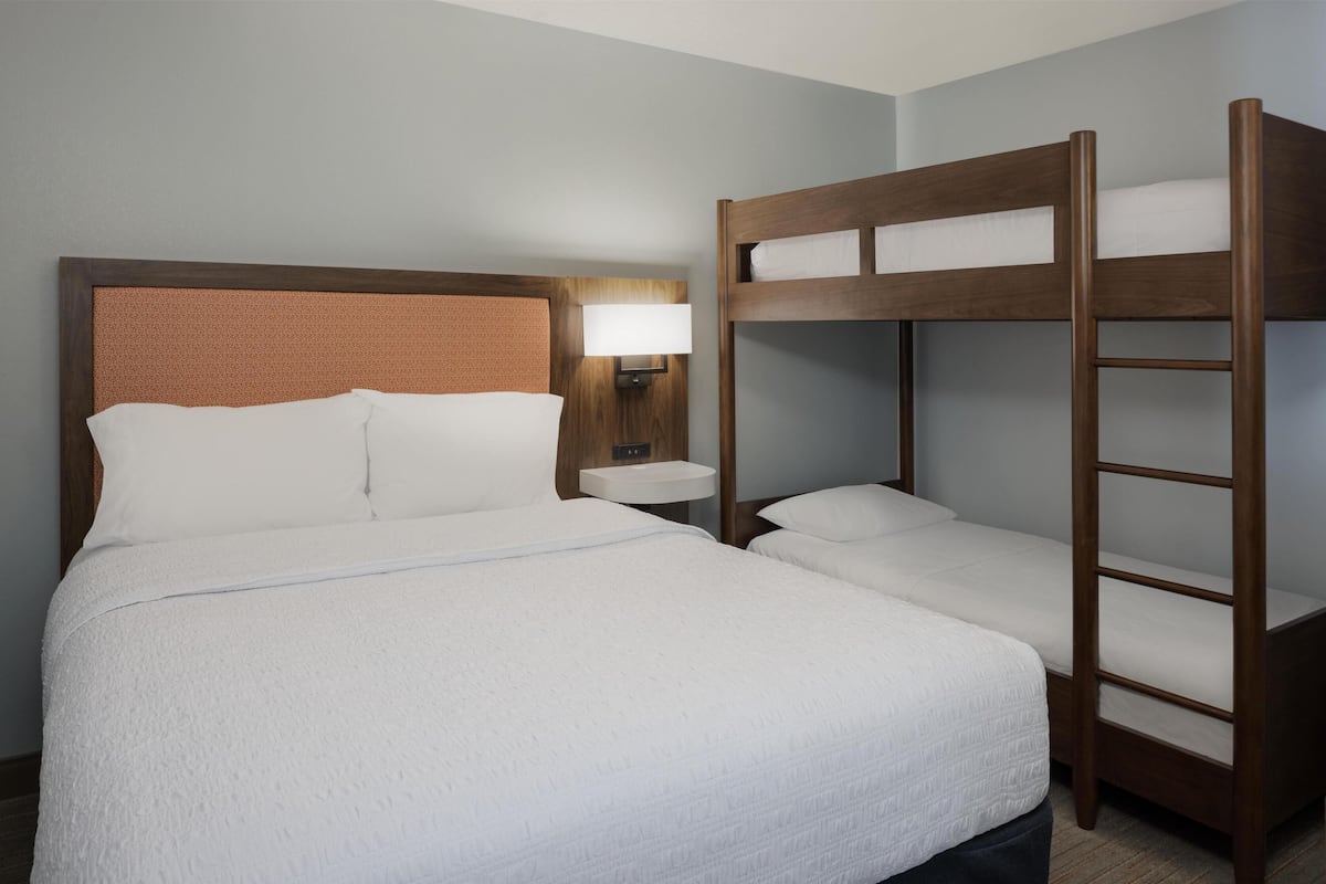 A hotel room with a large bed, a wooden bunk bed, a bedside table, and a wall-mounted lamp.