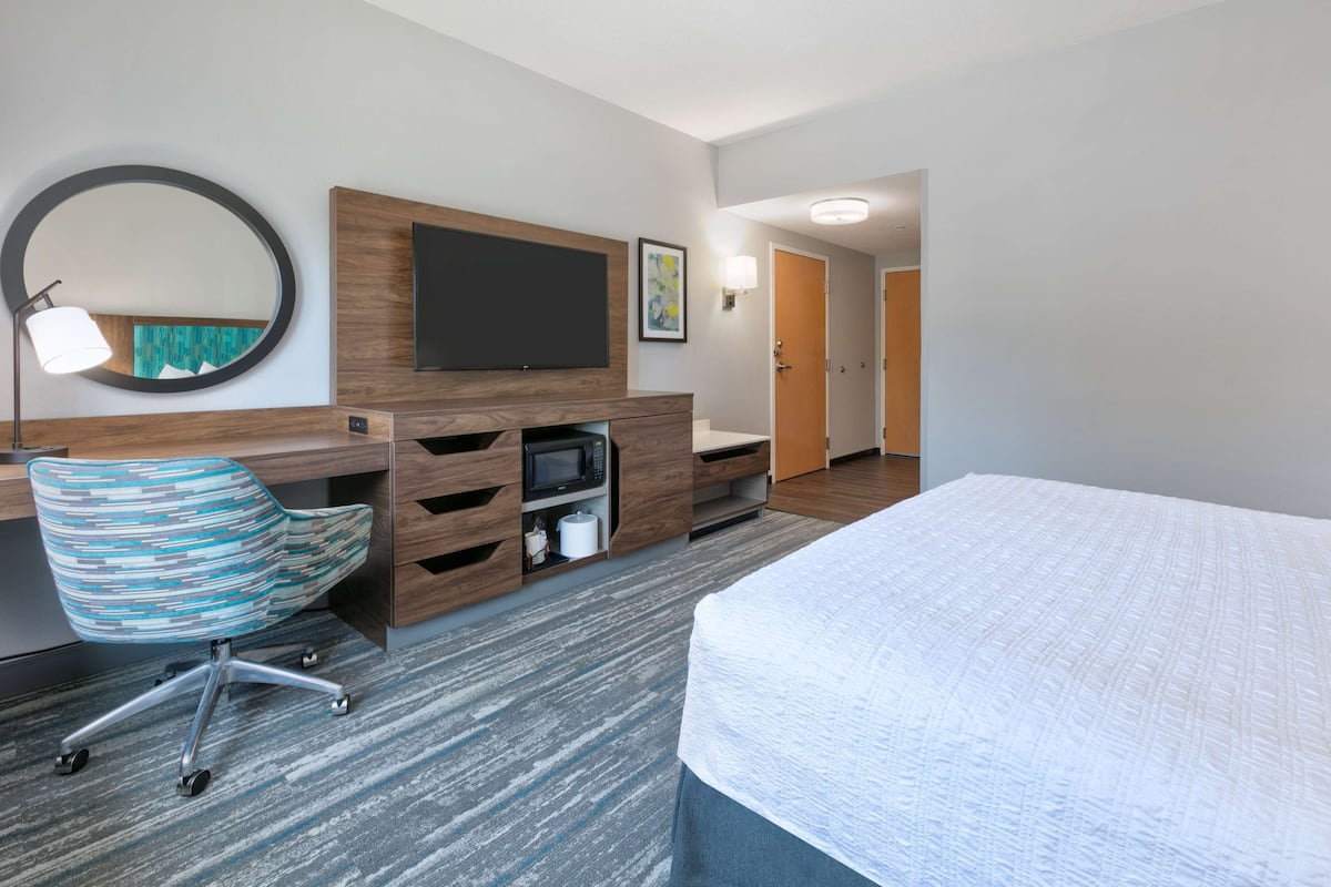 A hotel room with a bed, a desk with a flat-screen TV, a chair, a microwave, and a large mirror.