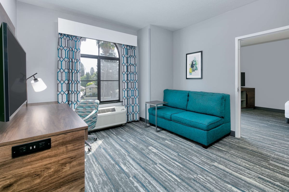 A modern living room with a teal sofa, a wooden desk, a standing lamp, and a large window with patterned curtains.