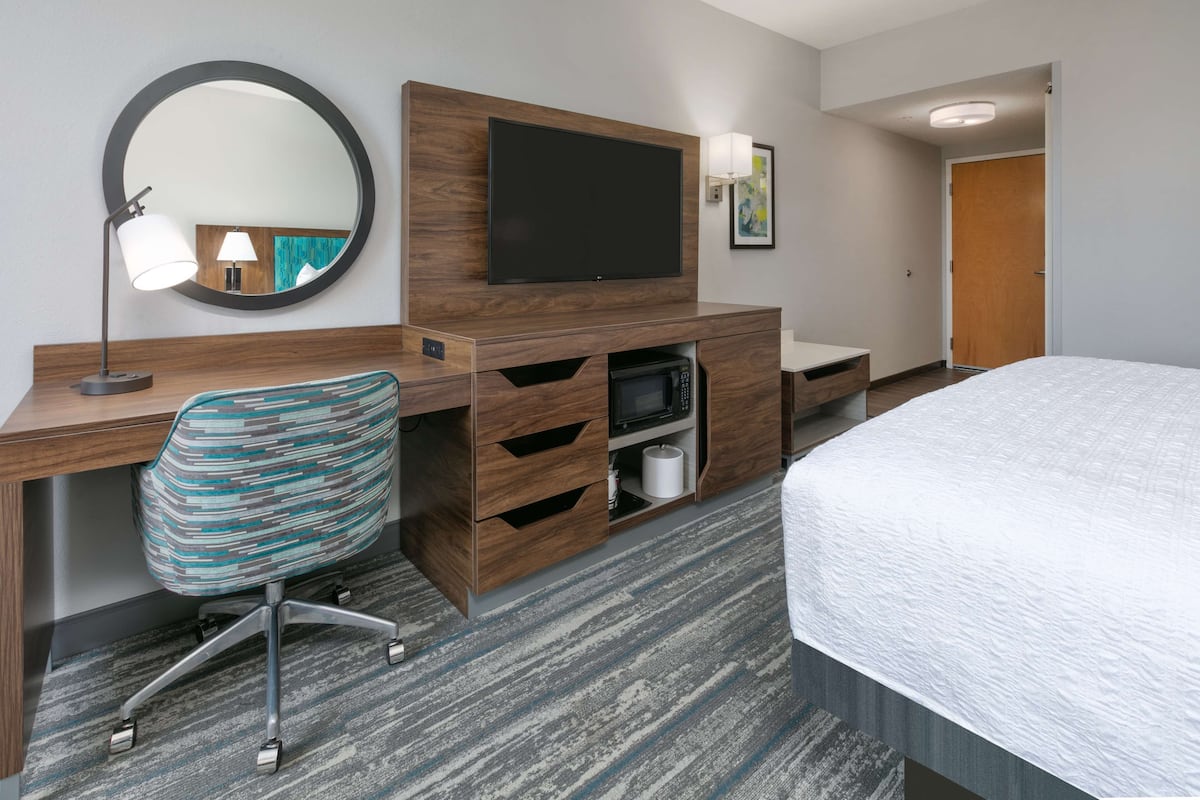 A hotel room with a wooden desk, a television, a mirror, a patterned chair, a bed, and a bathroom with a toilet and sink.