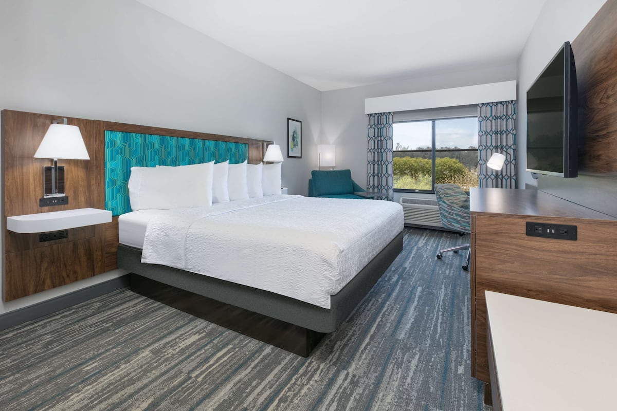 A modern hotel room with a large bed, a desk, a TV, and a window with a view of the outdoors.