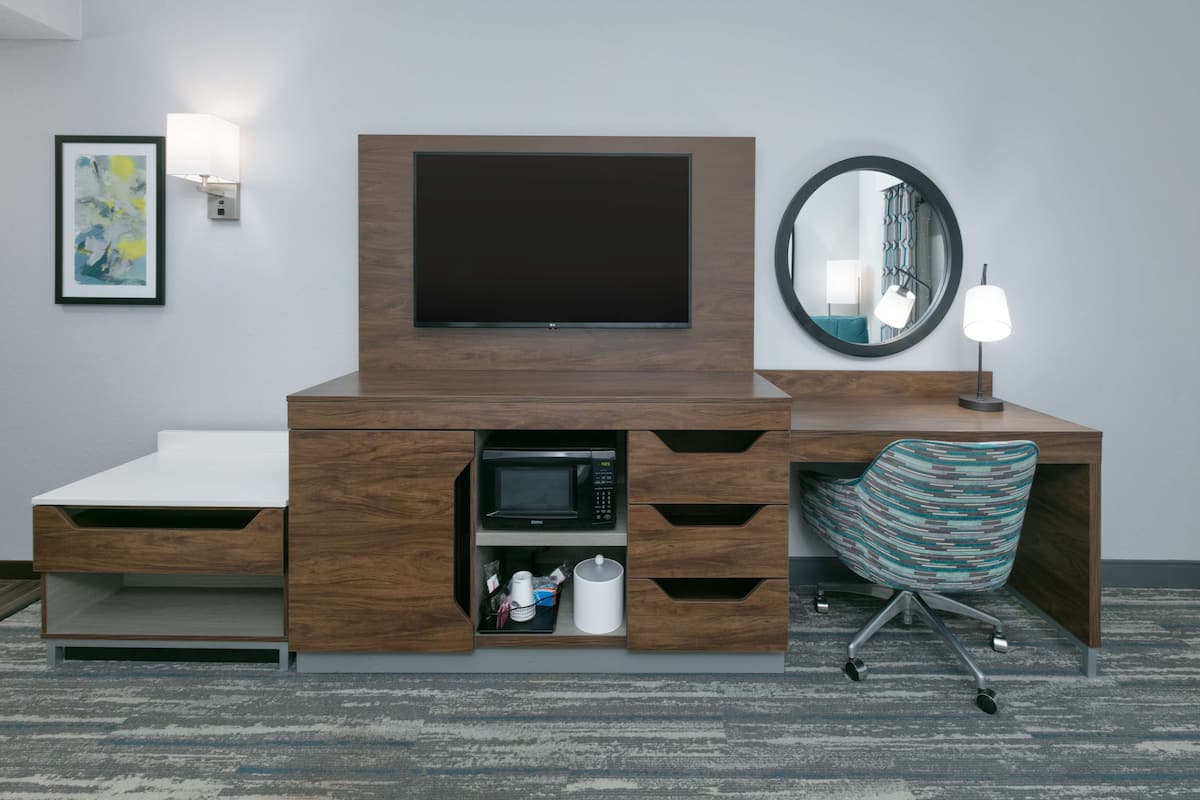 A modern hotel room with a wooden desk, a flat-screen TV, a microwave, a coffee maker, and a striped chair.