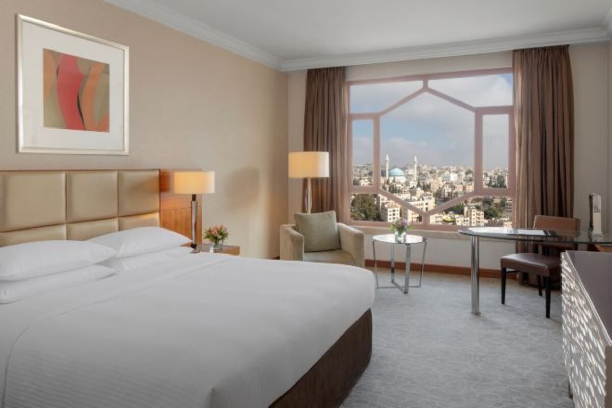 A hotel room with a large bed, a desk, a chair, a small table, and a window with a city view.