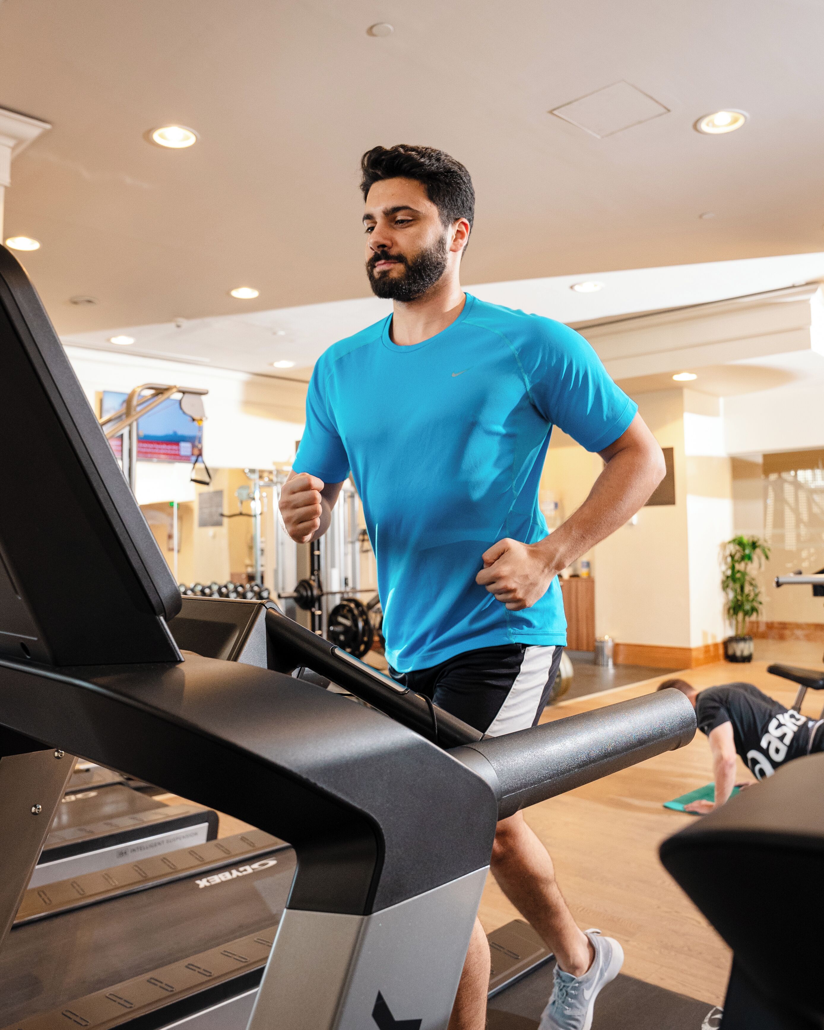 Un homme marche sur un tapis de course dans une salle de sport, avec des appareils d’exercice et un banc de musculation en arrière-plan.