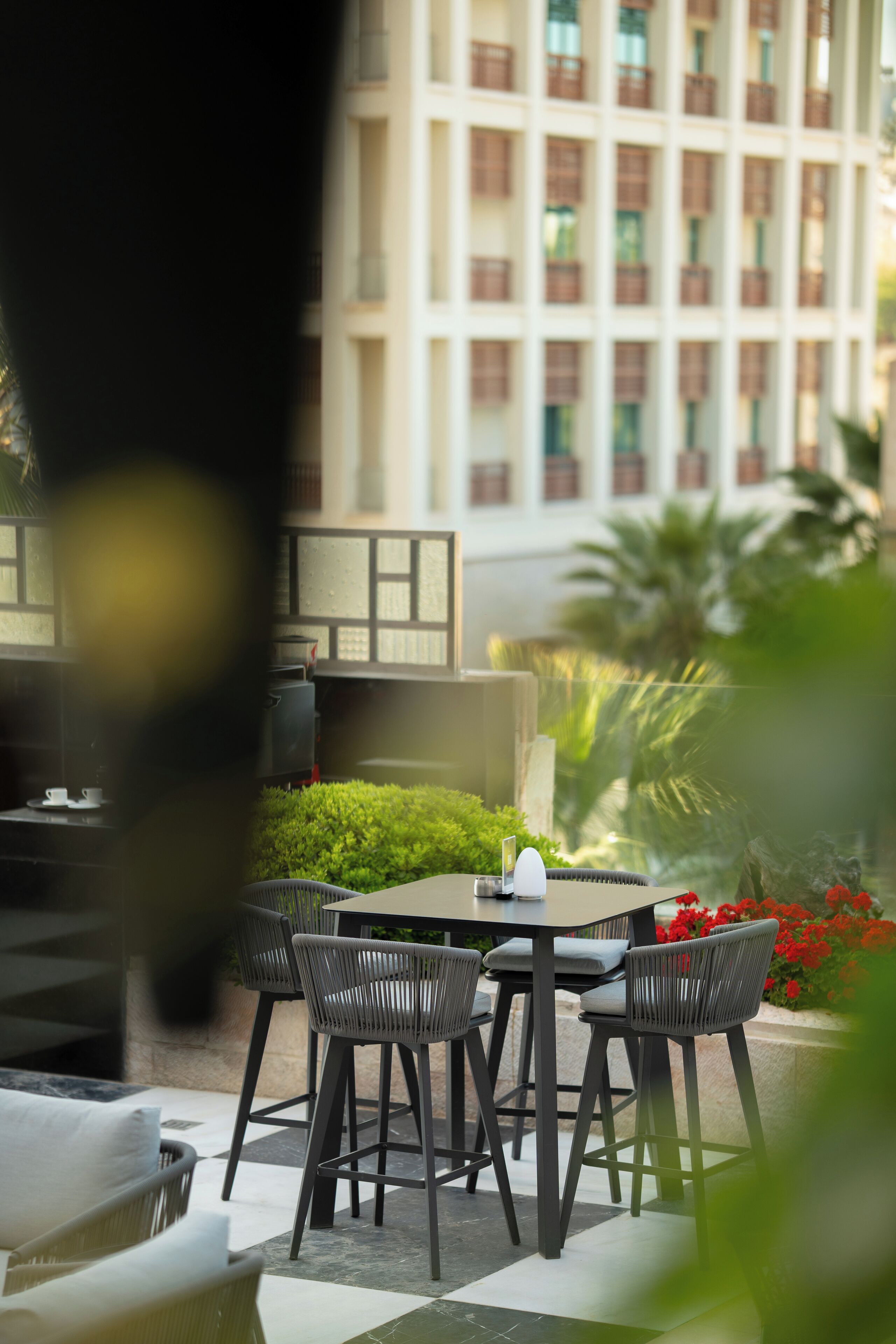 An outdoor seating area with a table and chairs, surrounded by greenery and a building in the background.