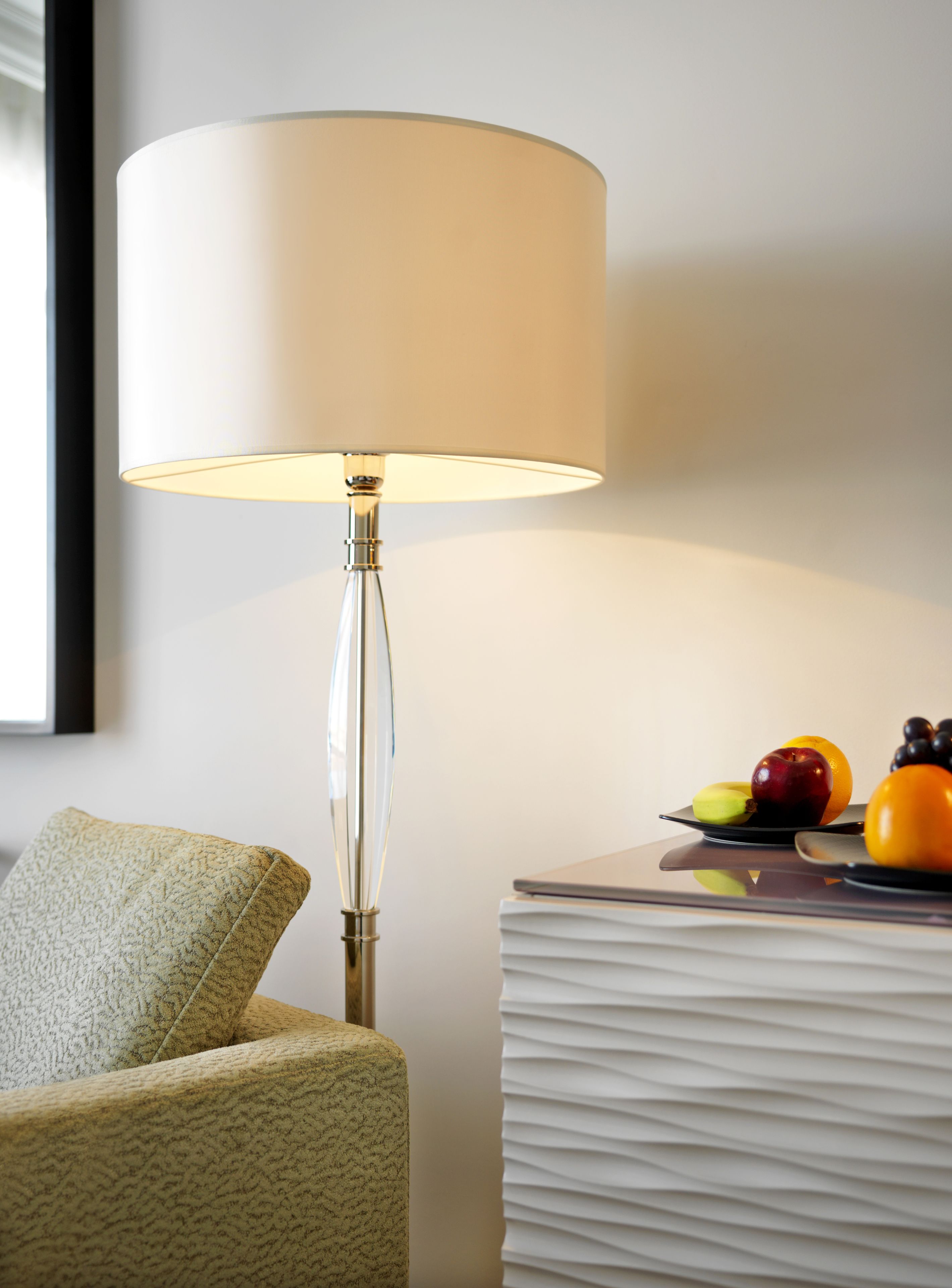 A modern lamp with a clear shade, a textured armchair, and a fruit plate on a white side table.