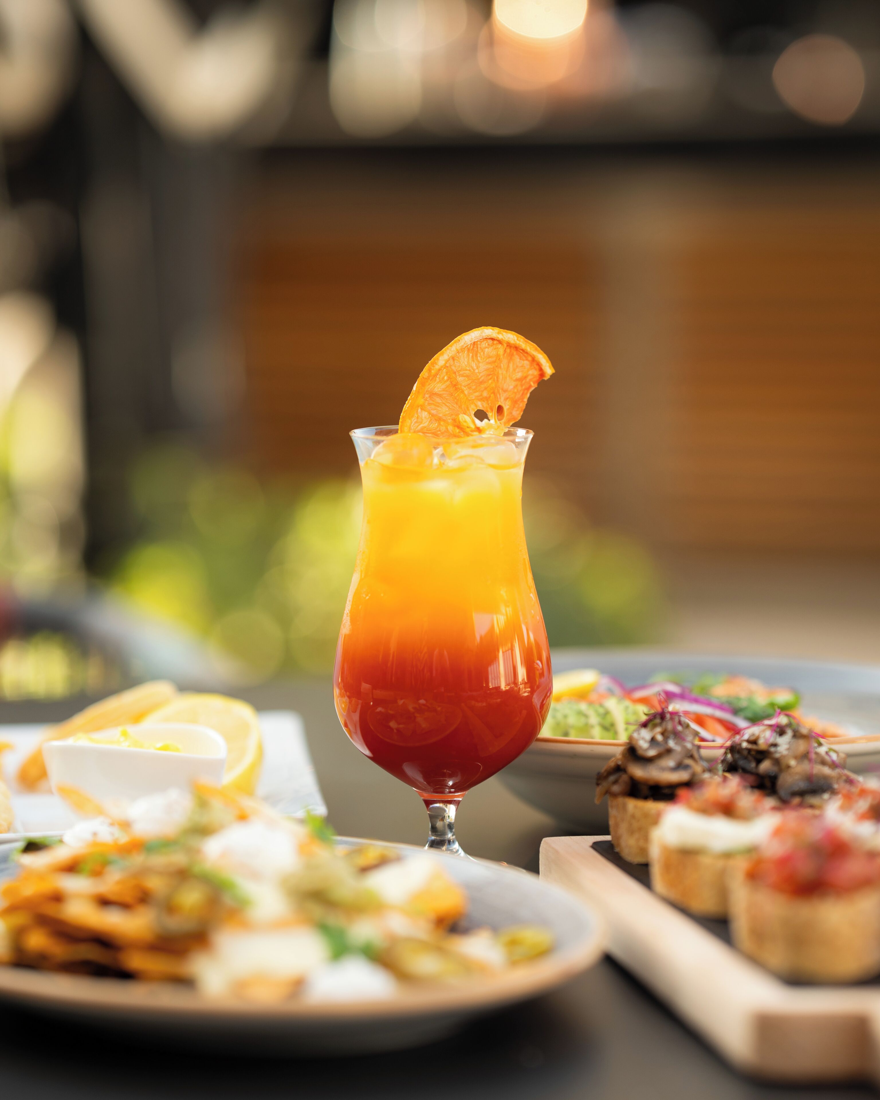 A glass of orange juice with a slice of orange on the rim, a plate of food, and a wooden board with appetizers.