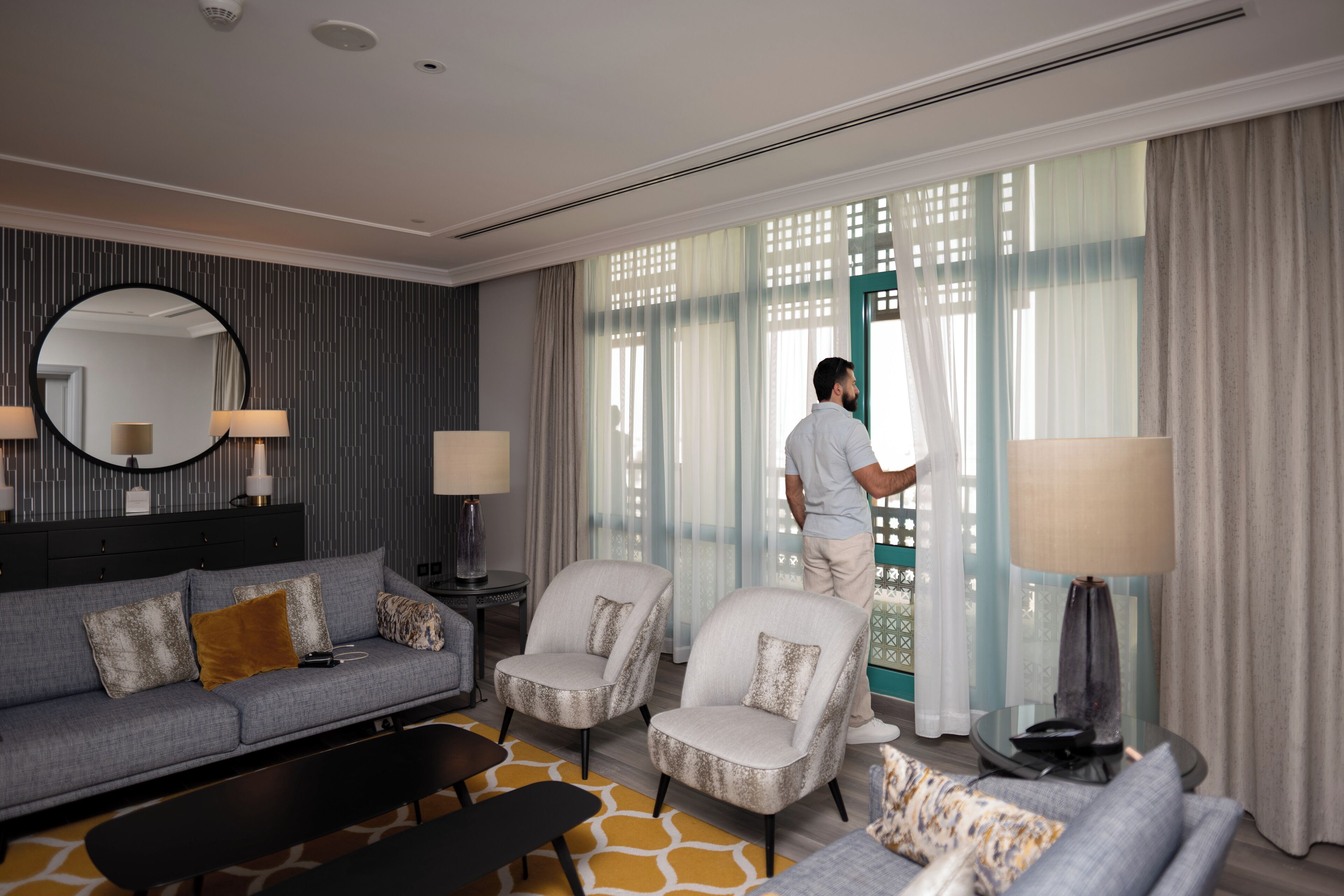 A modern hotel room with a grey sofa, two armchairs, a round mirror, and a large window with sheer curtains.