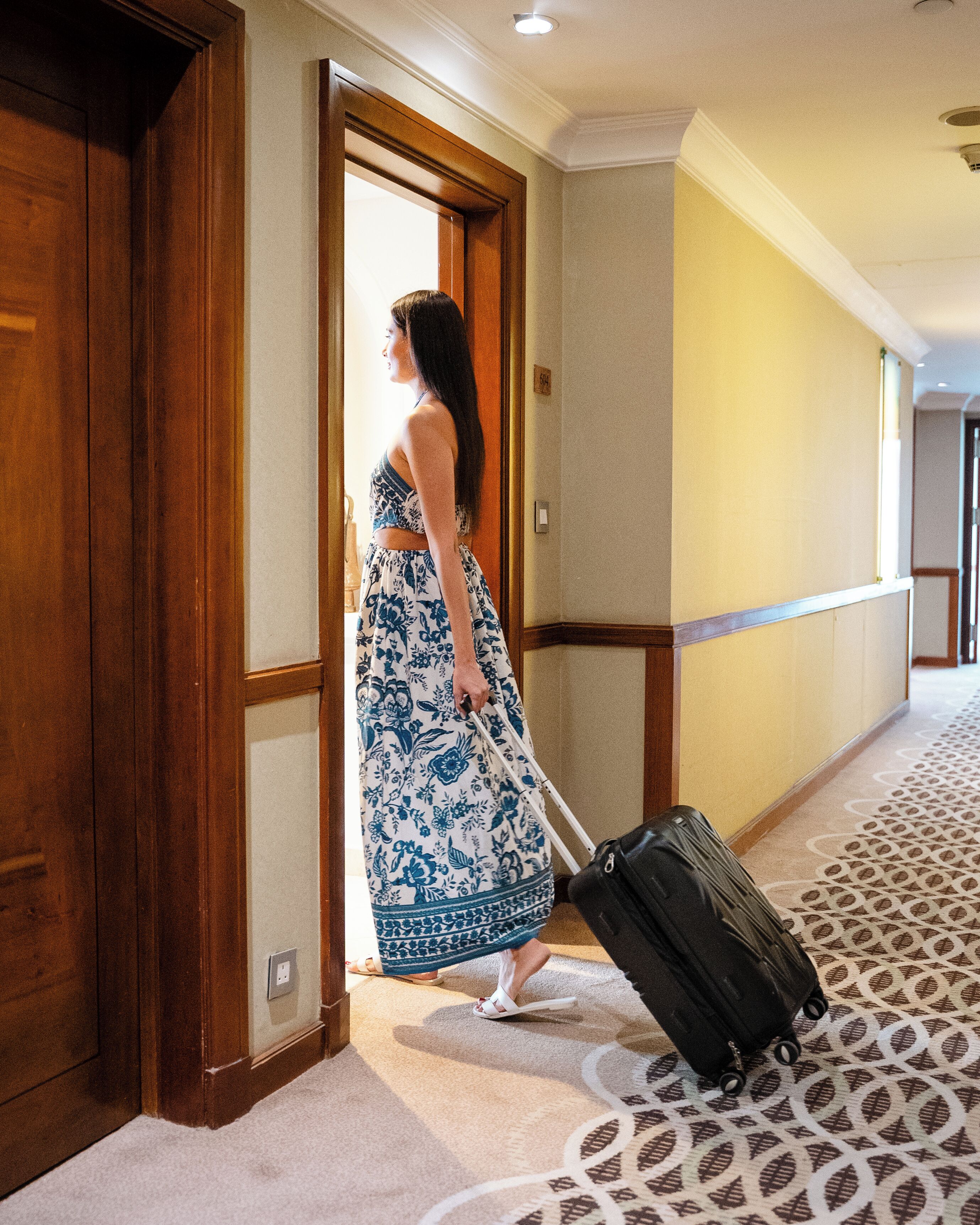 A woman in a floral dress exiting a hotel room, with a suitcase on the floor.