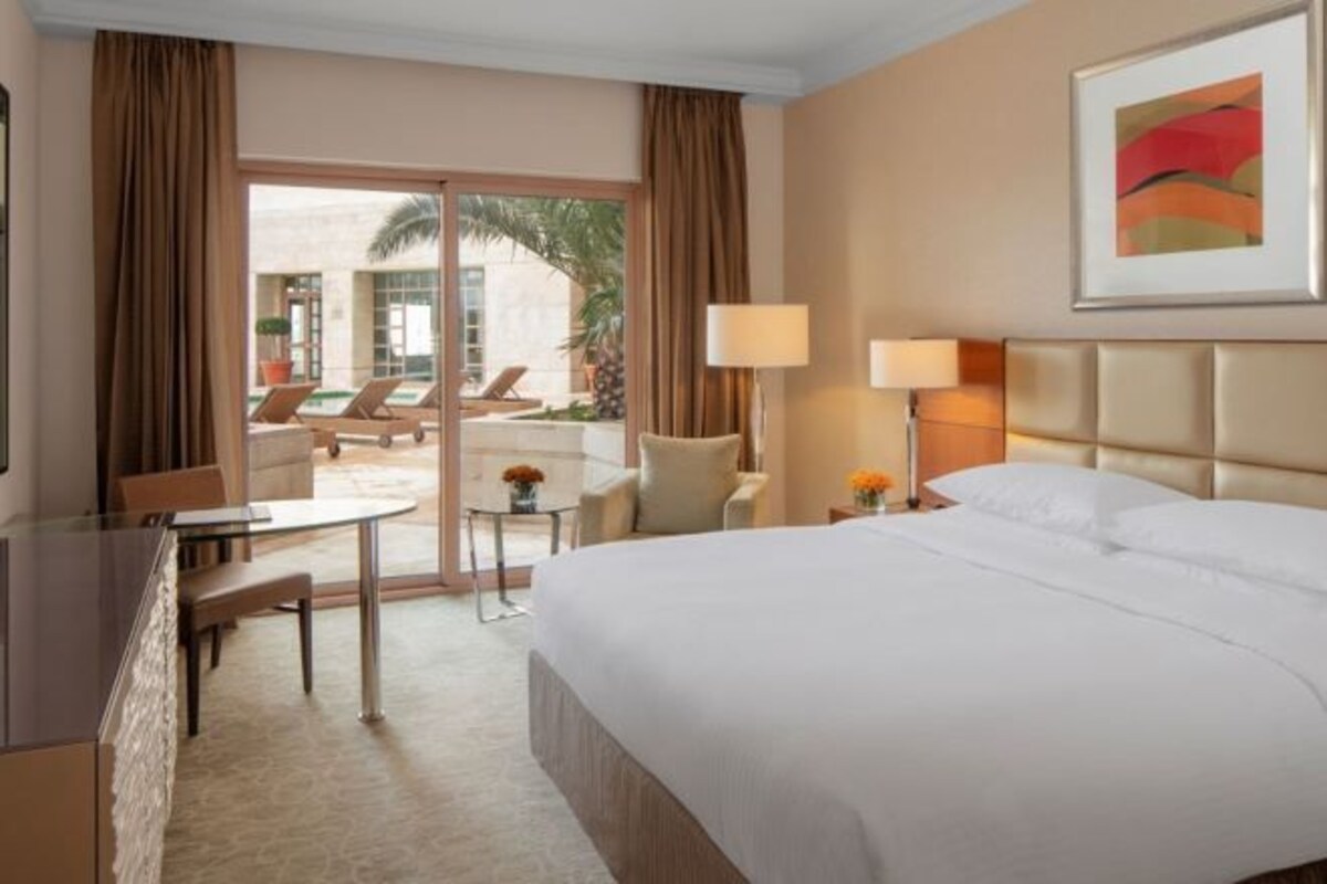 A hotel room with a bed, a desk, a chair, a lamp, and a view of the outdoor area.