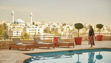 A rooftop pool with lounge chairs, potted plants, and a cityscape view in the background.
